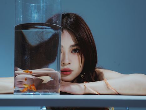 A creative portrait of a woman, partially obscured by a clear glass vase containing goldfish, shot indoors.